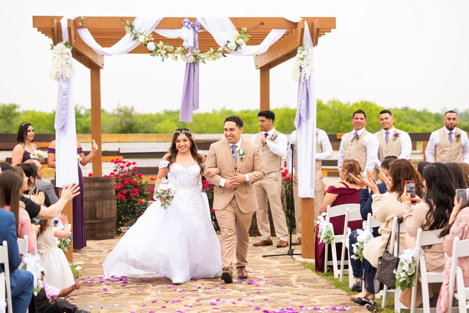 A smiling newlywed couple walks down the flower-lined aisle as guests applaud