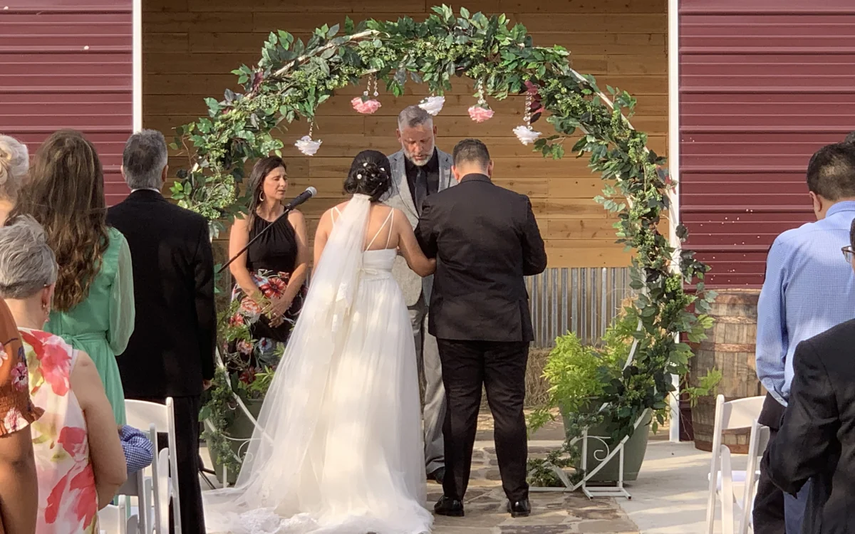 A couple exchanges vows beneath a leafy outdoor arch surrounded by guests